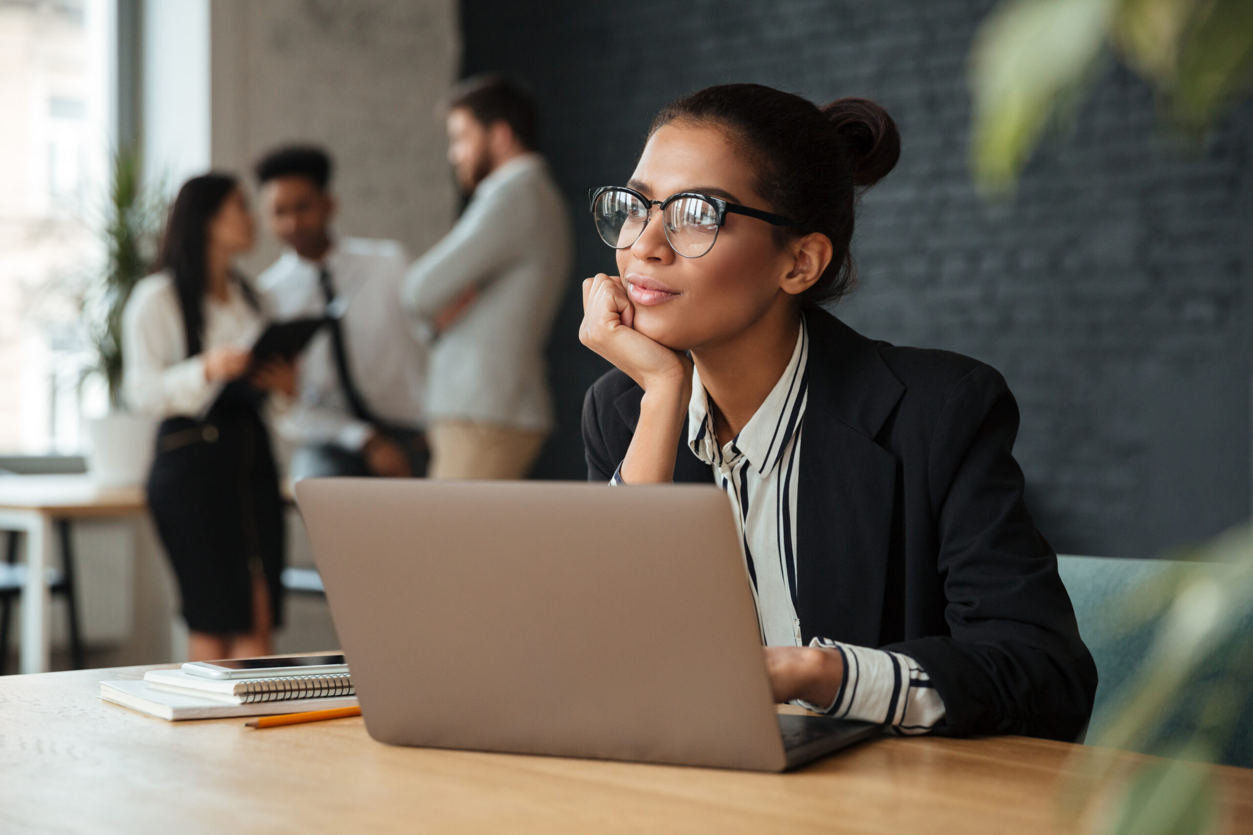 Photo of dreaming young african business woman sitting indoors using laptop computer. Looking aside.