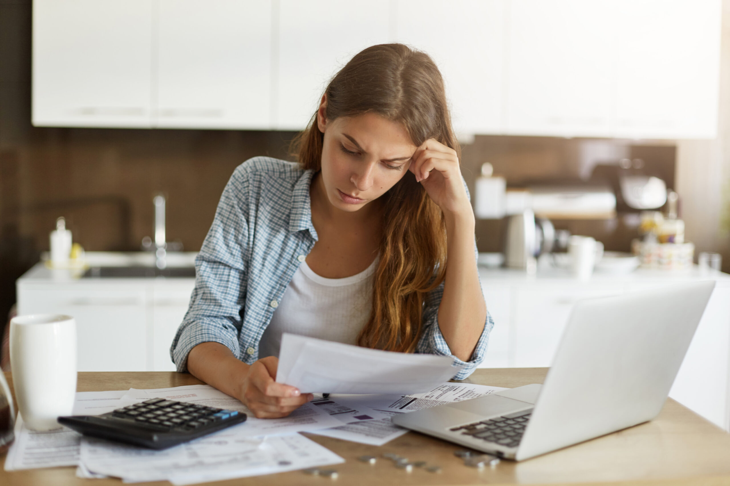 Attractive young housewife wearing shirt at home studying gas and electricity bills, checking calculations, looking at sheet of paper in her hands with serious and focused expression on her face